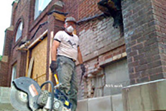 A worker wearing a hat and a mask is working beside a building.
