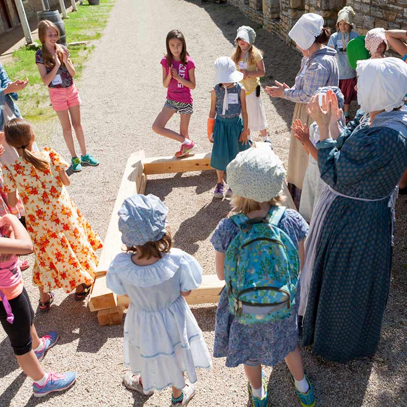 A group of young girls in bonnets gather around a costumed camp guide on a gravel path.