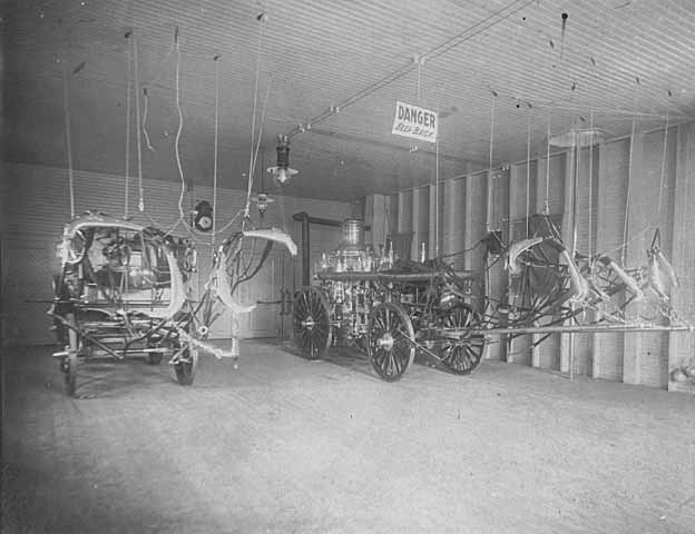 Photo of fire station interior showing horse-drawn pumper and wagon for equipment, ca. 1900.