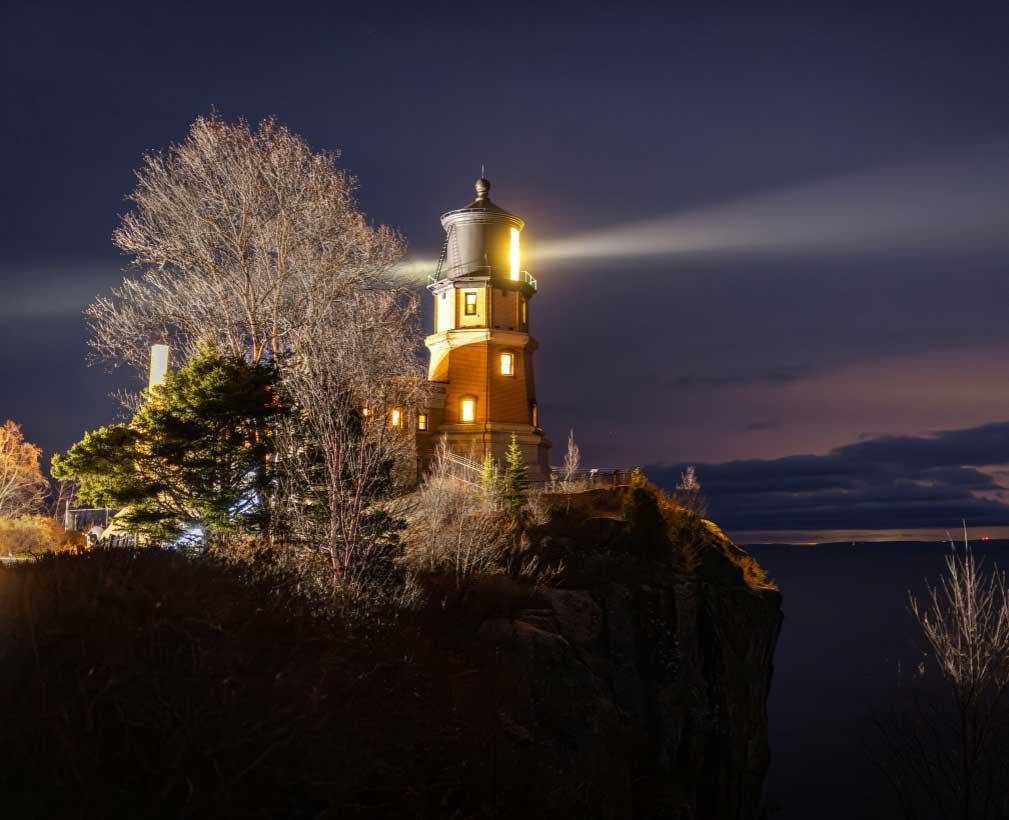 Image for the event Edmund Fitzgerald: A Great Lakes Memorial. At night, the historic Split Rock Lighthouse glows warmly atop a steep, dark cliff, casting a powerful, bright beam of light across the night sky and the waters of Lake Superior.