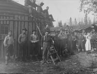 Detail of a panoramic photograph of lumberjacks in lumber camp, including men standing near a ladder propped against a building and others sitting on the building's roof.