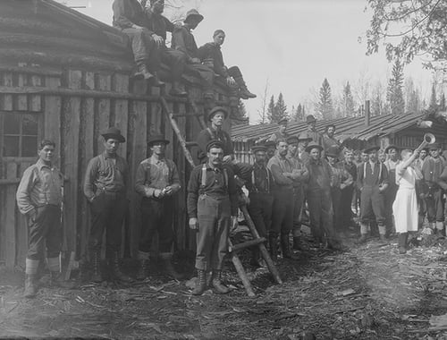Detail of a panoramic photograph of lumberjacks in lumber camp, including men standing near a ladder propped against a building and others sitting on the building's roof.