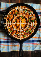 Lines of light-orange condiment arranged in the design of gun cross-hairs sit atop a hotdish in a black cast-iron skillet on top of a checkered cloth.