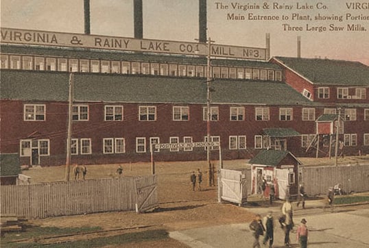 A four-story sawmill with a green roof, red walls, and pairs of windows across the horizontal access. People are in the foreground.