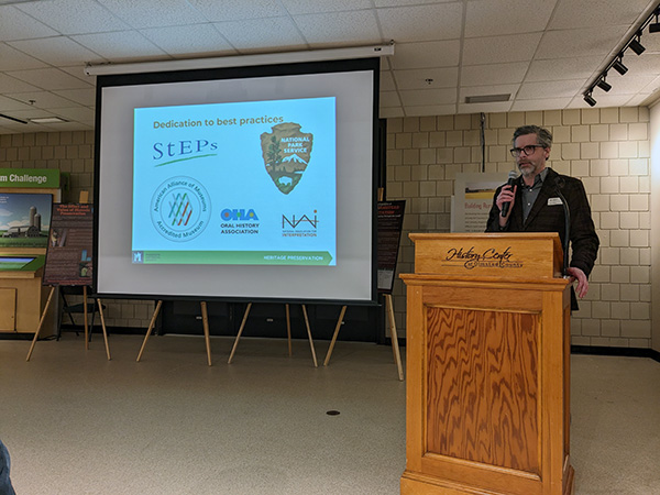 Todd Mahon presenting at podium with screen next to him showing PowerPoint presentation -1