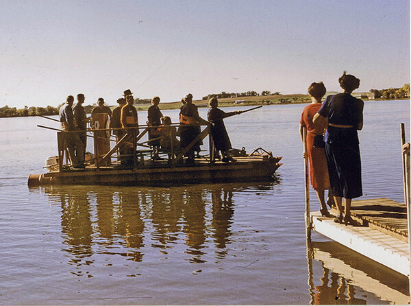 1959 color photo of group of 12 or so people standing on a pontoon boat in a lake with two women watching them from a dock