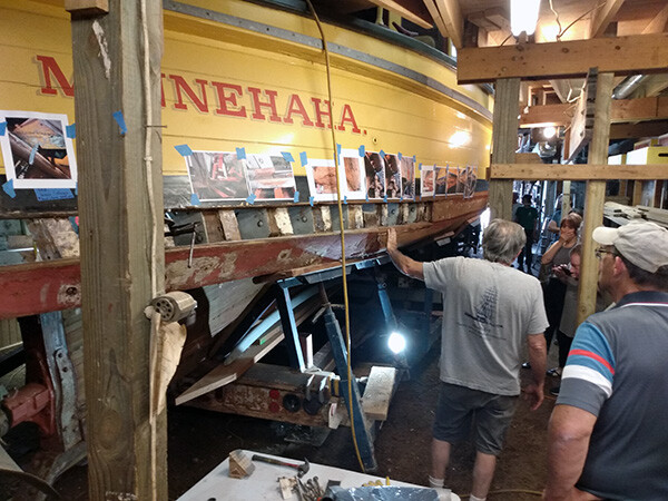 Steamboat Minnehaha on blocks with several people listening to a man talk about renovation work. Photos are pasted on the side of the boat