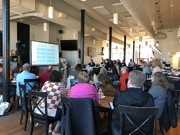 Presenter talking in front of crowd of people sitting at tables in a coffee shop-Main Street Conference Owatonna
