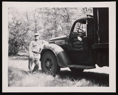 Man leaning against front of truck. Text on door of truck says, Minnesota Department of Conservation, State Parks-ca. 1940
