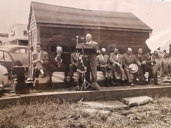 Man speaking at podium draped with banner that reads Radio Station WKLK. Behind him are 8 men sitting in a row on a bench. Behind them is a wood shed. Year 1950