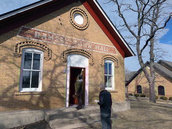 Front exterior of the Ramsey Town Hall building with one person in the open doorway pointing to the top of the doorframe and another person stands in the foreground