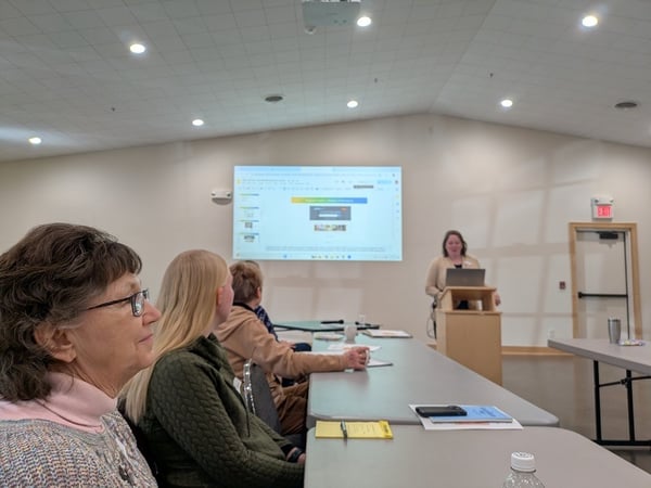 On the left side of photo are participants listing to a speaker at a podium with powerpoint presentation behind her
