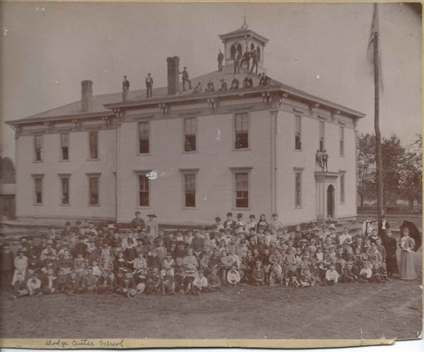 Many young people all over the roof, in windows, on cupola of school building. Large numbers of children in the yard. The flag pole is of a tree