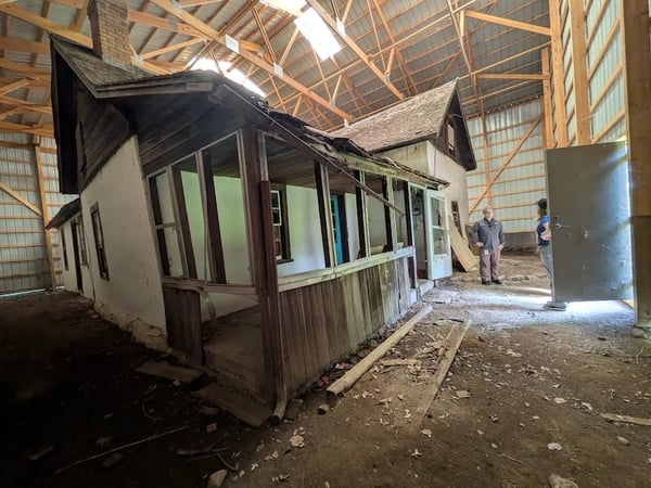 _Interior shot showing a metal building surrounding an old farm house. Two people  are inside the metal building talking copy
