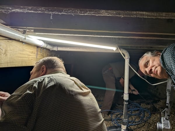 Three people investigatign cramped crawl space that is dimly lit by a improvised light fixture 