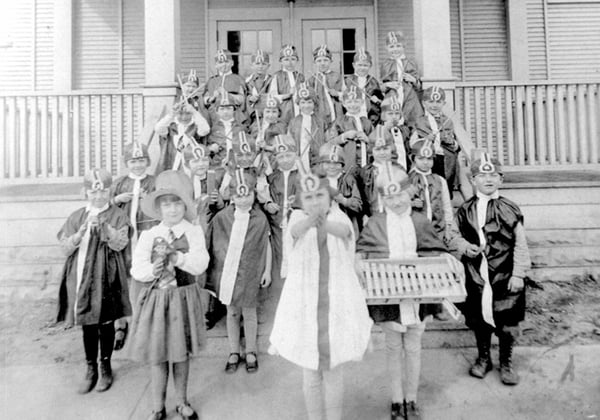 Group of around 25 children standing on steps school poarch in festive costumes holding sticks. One child has a xylophone
