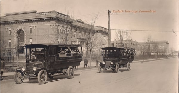 Two school buses with kids in the 1920s with school buildings behind them