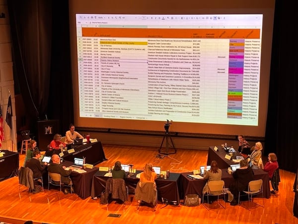 Meeting with 11 people set up on a stage. On large screen is a spreadsheet
