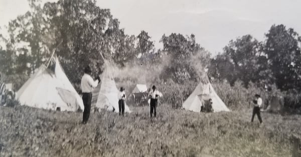 Blurry photo. Four people possibily White Earth Ojibwe possibily  playing baseball in front of three tipis. Two children are coming out of one tipi