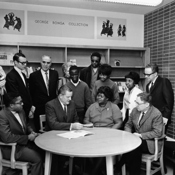 Group of African American students and African American and white staff standing and sitting around a table listening to someone talking