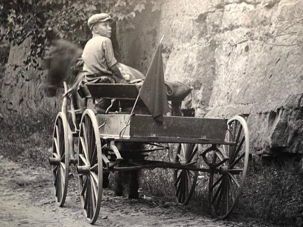 Boy sitting in open wagon pulled by horse. Wagon is by a rocky wall
