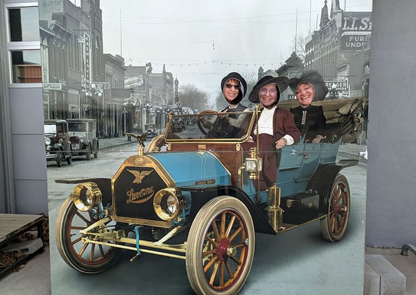 image depicts a face-in-the-hole board featuring a scene with three people drive an antique car
