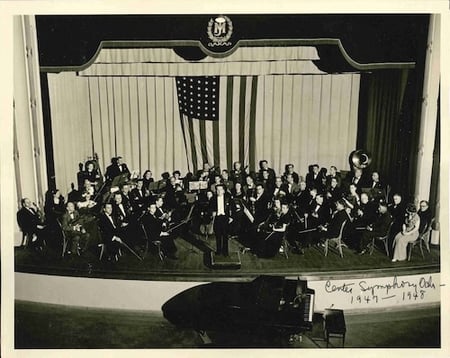 Black and white photo of the Center Symphony Orchestraon stage with a conductor and an American flag backdrop. 1947-48