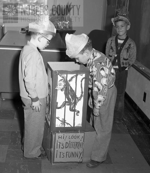 Three boys in a classroom wearing paper hats, with two observing a box painted with a monkey.
