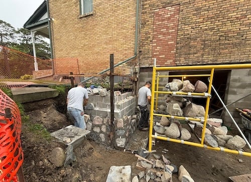 Two workers constructing a stone structure beside a brick building with scaffolding and rocks