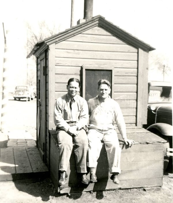 Two men seated on a wooden structure in front of a small shed, with vintage cars in the background