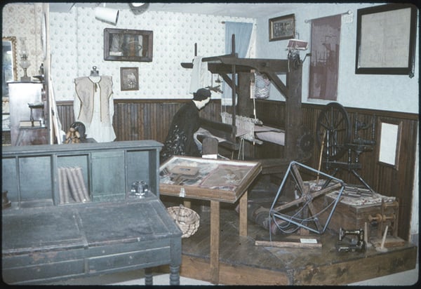 A museum exhibit room with an old wooden loom, spinning wheel, mannequin in period dress, and vintage textile tools