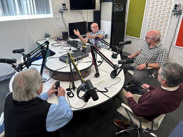 Four men seated around a radio studio table with microphones and equipment.