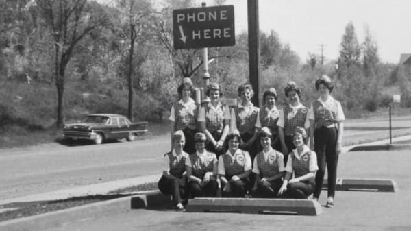 A group of women in uniform poses in front of a PHONE HERE sign, with trees and a vintage car in the background
