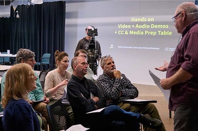A workshop on media archiving with attendees engaged in discussion. An instructor stands at the front, gesturing, with a camera in the background and a screen displaying workshop details.