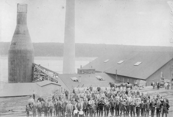 A historic black-and-white photo of the Schulenburg Lumber Mill circa 1885, showing a group of people in front of large industrial structures, including a kiln and smokestack, amidst a rural landscape.