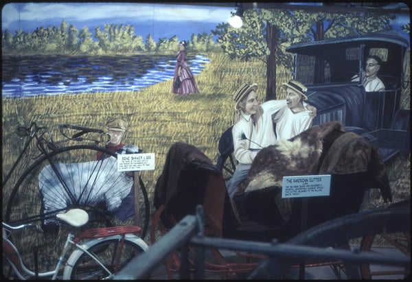 A painted exhibit mural showing a rural scene with a bicycle, two men in a horse-drawn carriage, and a woman in a dress by the water