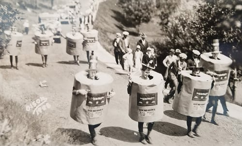 A 1928 4th of July parade showing participants in large beige cylindrical costumes for "Balsam Wool," walking along a gravel road near Pinehurst Park, with onlookers in the background.