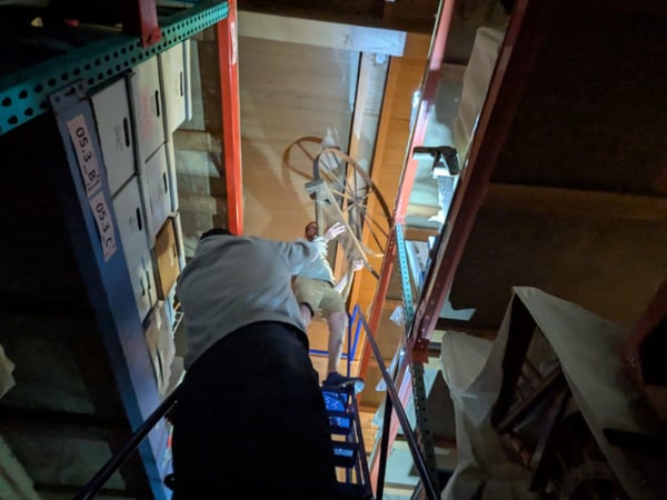 Two individuals in an archive are passing a spinning wheel down a ladder; one is on the ladder while the other is above handing it down