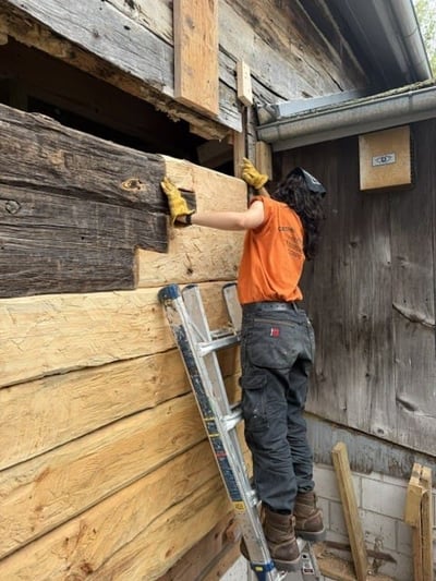 A person on a ladder working on a wooden wall, wearing an orange shirt and gloves, with tools scattered at the base of the ladder
