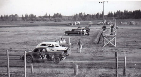 Black and white photo of two cars racing on dirt track