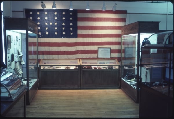 Interior of an exhibition space featuring a large American flag backdrop with display cases containing various artifacts in the foreground.