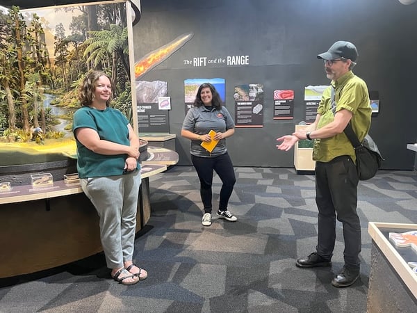 Three people converse in a museum exhibit featuring a diorama of a lush landscape. Informational panels are displayed in the background, including one that reads "The RIFT and the RANGE."