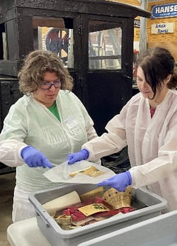 Two women in protective clothing working together at a table, examining artifacts in a gray container in a workshop or museum setting.
