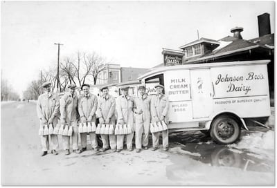 Eight male dairy workers in overalls stand outside a Johnson Bros. Dairy truck, each holding several bottles of milk.