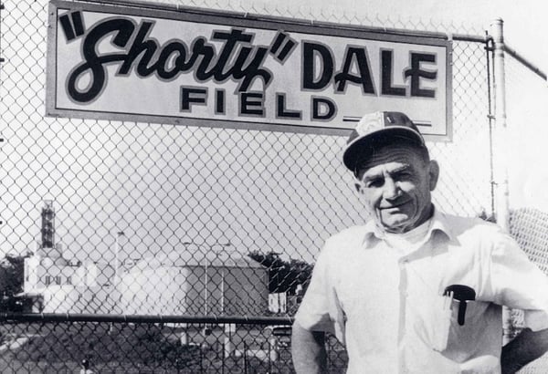 A older man in baseball hat standing in front of chain link fence with a sign that says, "Shorty" Dale Field