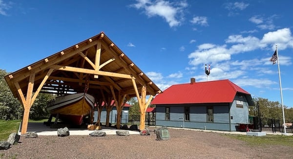 A yellow and red boat named "LEIF ERIKSON" sits under a wooden shelter at a historical site, with a gray building and American flag visible nearby against a blue sky.