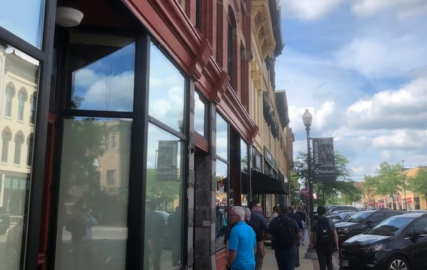 Touring a downtown street featuring glass storefronts, pedestrians, parked cars, and a "Welcome" banner.