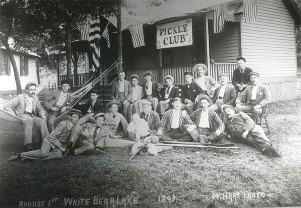 Black and white photograph of a group of men posing outside the Pickle Club building at White Bear Lake in 1893, surrounded by greenery and American flags