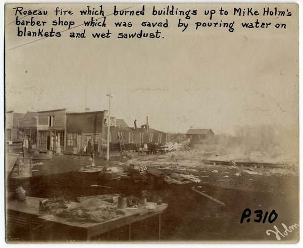 Black and white photo of burnt smoldering buildings with text Roseau Fire which burned buildings up to Mike Holm's barber shop which was saved by pouring water on blankets and wet sawdust.