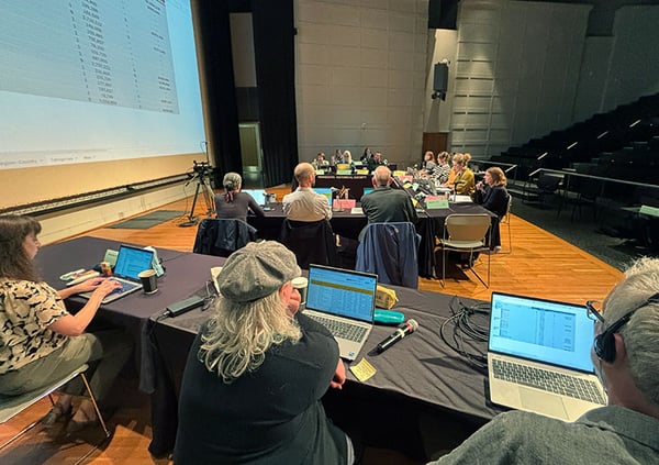 Staff work on laptops with HRAC members around tables during a meeting in a tiered lecture hall, with a spreadsheet projected on a large screen.